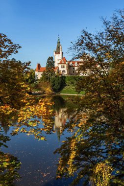 Pruhonice, Çek Cumhuriyeti - 25 Ekim 2020: Sarı ve portakal ağaçlarıyla çevrili bir parkta duran romantik şatonun manzarası. Şatonun suya yansıması. Güneşli sonbahar günü, mavi gökyüzü.