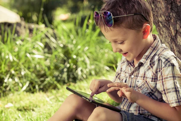 Glückliches Kind mit Tablet-PC, das tagsüber neben einem Baum sitzt. — Stockfoto