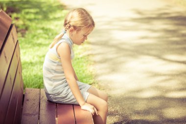 sad little girl sitting on bench in the park at the day time clipart