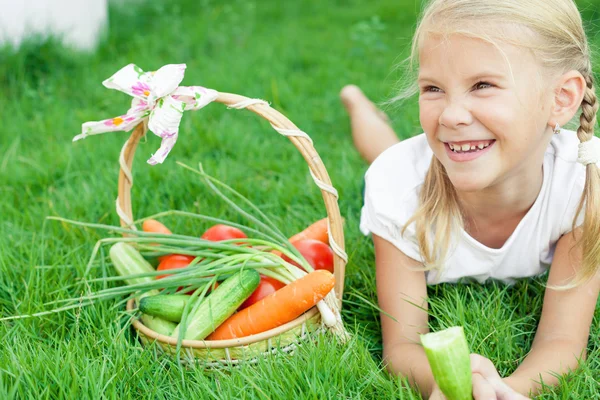 Happy little girl lying on the grass with a basket of vegetables — Stock Photo, Image