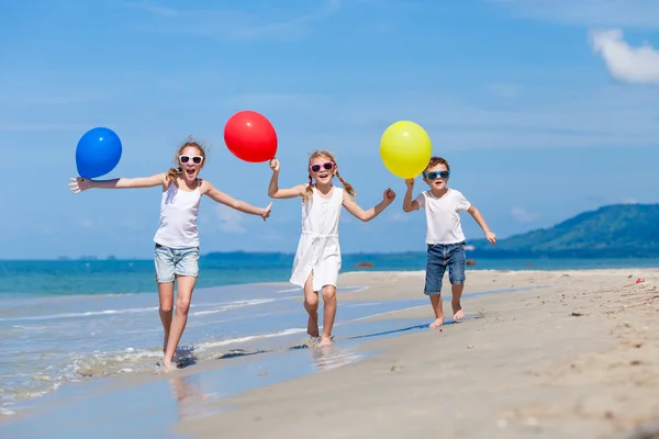Tres niños felices con globos corriendo en la playa en el d — Foto de Stock