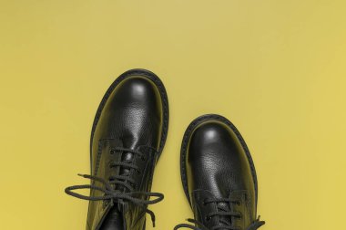 Sarı arka planda şık siyah deri çizmeler düz manzarada yatıyordu. Tarz sahibi kadınların erkek üniformaları. Ayakkabı bağcıklı Grunge çizmeleri. Kıyafetler, ayakkabılar, moda geçmişi. Klasik deri ayakkabılar..