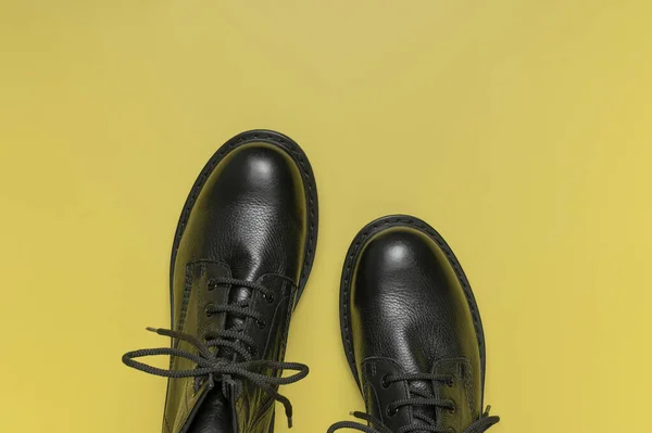 Fashionable youth black leather boots on yellow background flat lay top view. Stylish womens mens unisex boots. Grunge boots with bootlace. Clothes, shoes, fashion background. Classic leather shoes.