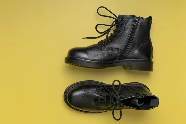 Fashionable youth black leather boots on yellow background flat lay top view. Stylish womens mens unisex boots. Grunge boots with bootlace. Clothes, shoes, fashion background. Classic leather shoes.