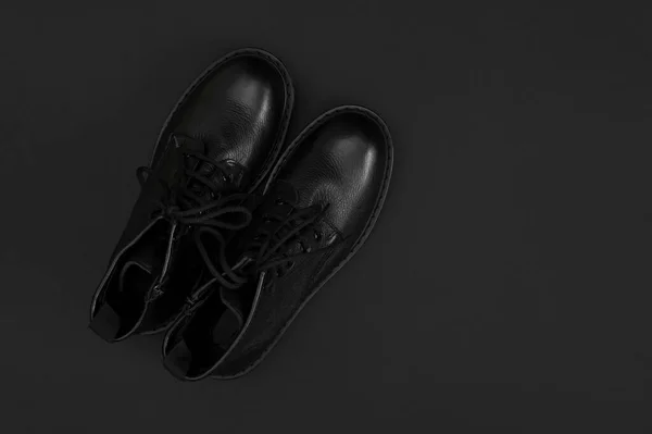 Fashionable youth black leather boots on black background flat lay top view. Stylish womens mens unisex boots. Grunge boots with bootlace. Clothes, shoes, fashion background. Classic leather shoes.