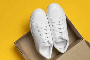 Beyaz kadınların deri spor ayakkabıları sarı arka plan manzaralı karton kutunun içinde düz yerde yatıyordu. Şık gençlik ayakkabıları, spor ayakkabıları, hakiki deri ayakkabılar. Minimalist ayakkabı mağazası reklamı, moda
