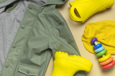Bebek sarı lastik çizmeler, yeşil kapüşonlu ceket, tahta oyuncak piramit, bej arka planda sarı şapka. Su geçirmez polimerik ayakkabılar. Yağmur ayakkabıları, çizmeler. Çocuk ayakkabıları Düz Sonbahar bahar bahar giysileri.