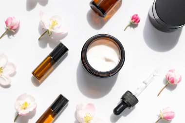 Doğal organik kozmetik bahar konsepti. Beyaz zemin üzerinde damlatan, kremalı, narin bahar çiçekleri olan temiz kahverengi cam kozmetik şişeleri düz bir zemin üzerinde yatıyordu. Güzellik bitkisi aromalı spa yağı..