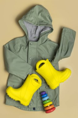 Bebek sarı lastik çizmeler, yeşil kapüşonlu ceket, tahta oyuncak piramit, bej arka planda sarı şapka. Su geçirmez polimerik ayakkabılar. Yağmur ayakkabıları, çizmeler. Çocuk ayakkabıları Düz Sonbahar bahar bahar giysileri.