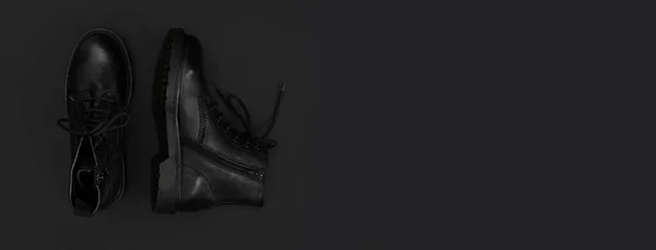 Fashionable youth black leather autumn boots on black background flat lay top view. Stylish womens mens unisex Grunge boots with bootlace. Clothes, shoes, fashion background. Classic leather shoes.