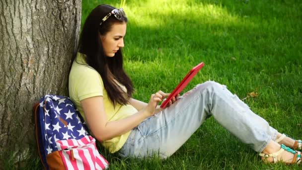Beeldmateriaal jonge mooie vrouw zitten in de buurt van een boom op het gras in het park en het gebruik van de app in de comuter Tablet PC — Stockvideo
