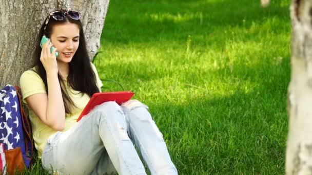 Filmaufnahmen junge schöne Frau sitzt neben einem Baum auf dem Gras im Park, spricht auf dem Smartphone und nutzt die App auf dem Tablet — Stockvideo