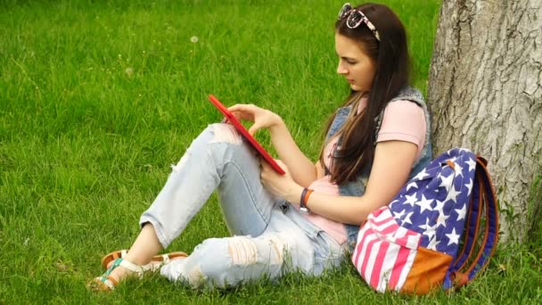 Materiał filmowy Młoda piękna kobieta siedzi w pobliżu drzewa na trawie w parku i korzystać z aplikacji w tablecie comuter. — Wideo stockowe