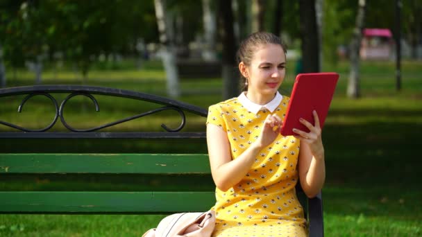 Mujer sentada en un banco en el parque y utilizar la aplicación en su tableta — Vídeos de Stock