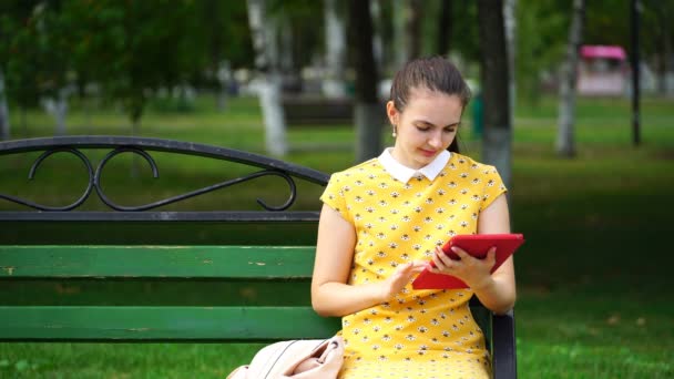 Mujer sentada en un banco en el parque y utilizar la aplicación en su tableta — Vídeos de Stock