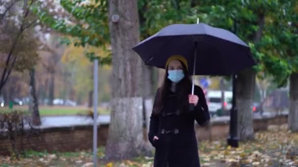 Uma jovem mulher com uma máscara protetora andando no parque sob guarda-chuva. Dia chuvoso, durante a segunda onda quarentena coronavírus COVID-19 pandemia — Vídeo de Stock