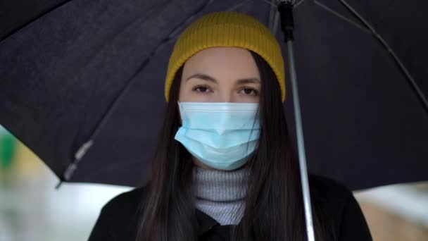 Een jonge vrouw met een beschermend masker die onder paraplu door het park loopt. Regendag, tijdens tweede golf quarantaine coronavirus COVID-19 pandemie — Stockvideo