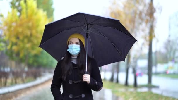 傘の下で公園を歩いている保護マスクの若い女性。雨の日,第二波隔離コロナウイルスCOVID-19パンデミック中 — ストック動画