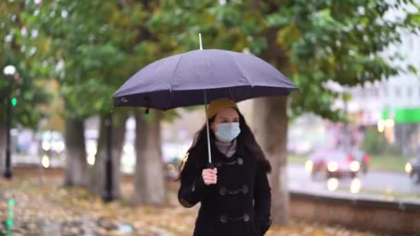 보호용 마스크를 쓴 젊은 여성이 우산 아래 공원을 걷고 있습니다. 비가 오는 날, 제2의 파도 검역 코로나 바이러스 COVID-19 유행병 — 비디오