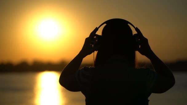 Žena poslouchá hudbu se sluchátky na pozadí západu slunce. — Stock video