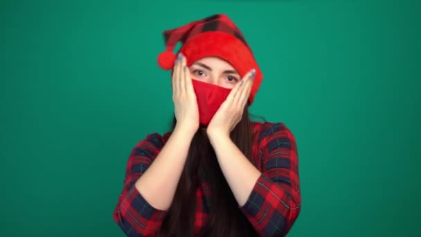 Ung kvinna i Santa hatt och röd medicinsk mask med chockade uttryck håller handflatorna på kinden — Stockvideo