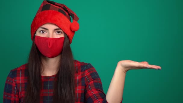 Young woman in santa hat and medical red mask holds invisible object, keeps palm raised — Vídeo de stock