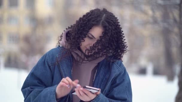 겨울 옷을 입고 겨울에 핸드폰을 사용하는 아름답고 곱슬머리의 젊은 여성 — 비디오