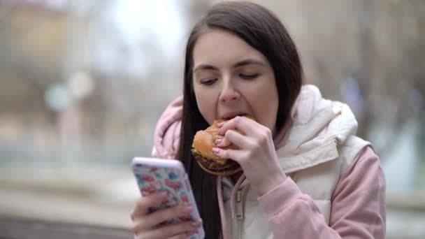 幸せな女性はハンバーガーを食べ、路上でスマートフォンを使用して — ストック動画