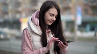 Cep telefonuyla yazan mutlu bir kadın şehir arka planında yürüyor.