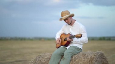 Hasır şapkalı bir adam samanlıkta gitar çalıyor.