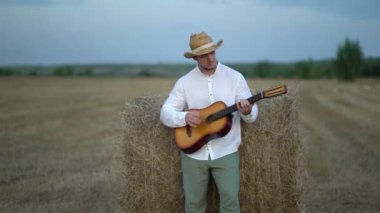 Hasır şapkalı bir adam samanlıkta gitar çalıyor.