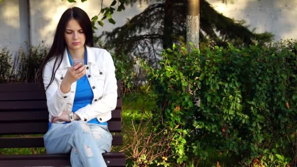 Chica joven utiliza la aplicación en su teléfono inteligente en el parque — Vídeo de stock