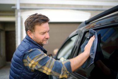 A mature man cleans his car with rag. Driver washes the glass of his car using microfiber washcloth. Father prepares the car for a family trip to nature clipart