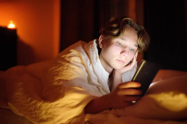 10代の男の子が電話でチャットしたり ブログに書いたり ソーシャルネットワークを読んで寝ています 若い男がベッドに横になり スマートフォンで音楽クリップを見ている ガジェットからの過剰使用と中毒の子供 — ストック写真