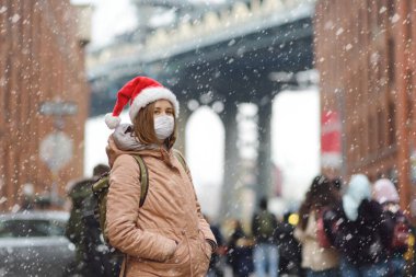 Noel Baba şapkalı genç bir kadın Noel arifesinde New York 'ta kar yağışı sırasında yürür. Yüz maskesi ve sosyal uzaklık - Coronavirus salgını sırasında, kış tatillerinde kamusal alanda güvenlik.