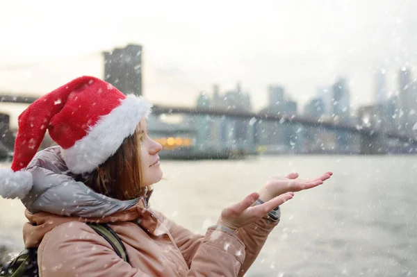 サンタクロースの帽子の地元の女性は 雪の多いクリスマス イブにニューヨークのブルックリン橋の近くの雪の中を歩いています 驚いた人は手のひらに雪の結晶をキャッチします ニューヨークの冬のクリスマスと新年の休日 — ストック写真