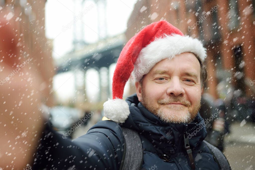 Local man in a Santa Claus hat takes selfie on street near the Manhattan Bridge in New York City on Christmas Eve during a snowfall. Winter Xmas holidays in NYC. New Year vacations in NYC.
