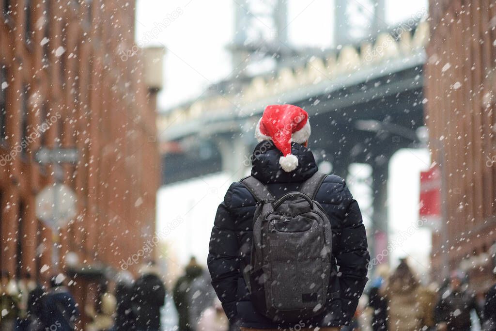 Middle aged man tourist in a Santa Claus hat is walking during a snowfall near Manhattan Bridge in New York on a snowy Christmas Eve. Winter Xmas holidays in NY. New Year vacations in NYC.