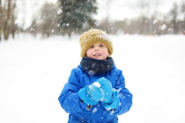 Küçük çocuk kar yağışında taze karla oynarken eğleniyor. Kartopu savaşı. Sıcak kıyafetler, şapka, eldiven ve eşarp giymiş bir çocuk. Karlı kış günlerinde doğada çocuk için açık hava eğlencesi.