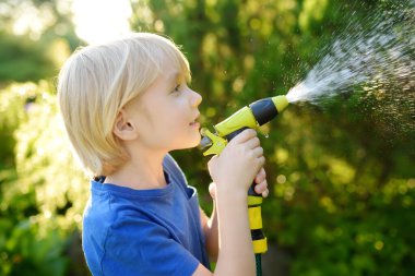 Çiçekleri sulayan, bahçe hortumuyla oynayan ve güneşli arka bahçede fıskiyeyle oynayan komik küçük çocuk. Anaokulu çocuğu su püskürterek eğleniyor. Çocuklar için yaz etkinliği.