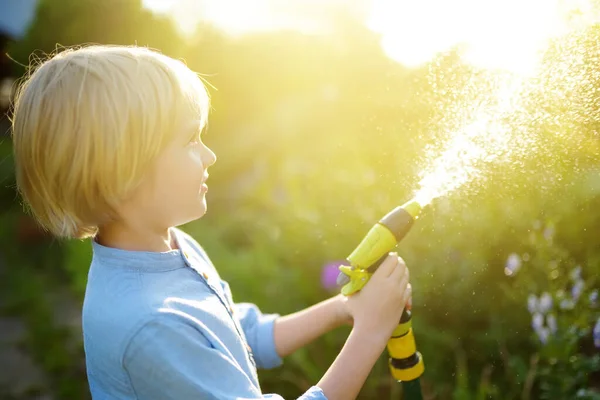 面白い小さな男の子は植物を散水し 日当たりの良い裏庭でスプリンクラーで庭のホースで遊んでいます 水のスプレーで楽しんでいる未就学児 子供のための夏の屋外活動 — ストック写真