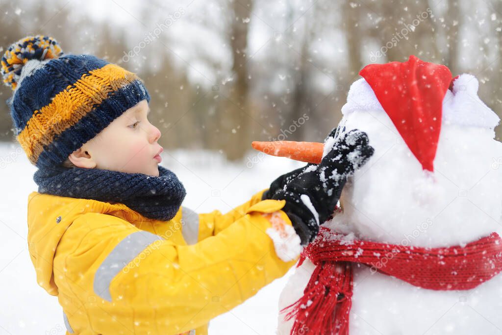 Little boy building funny snowman. ?hild attaches carrot nose to snowman in snowy park. Active outdoors leisure for children and family in winter