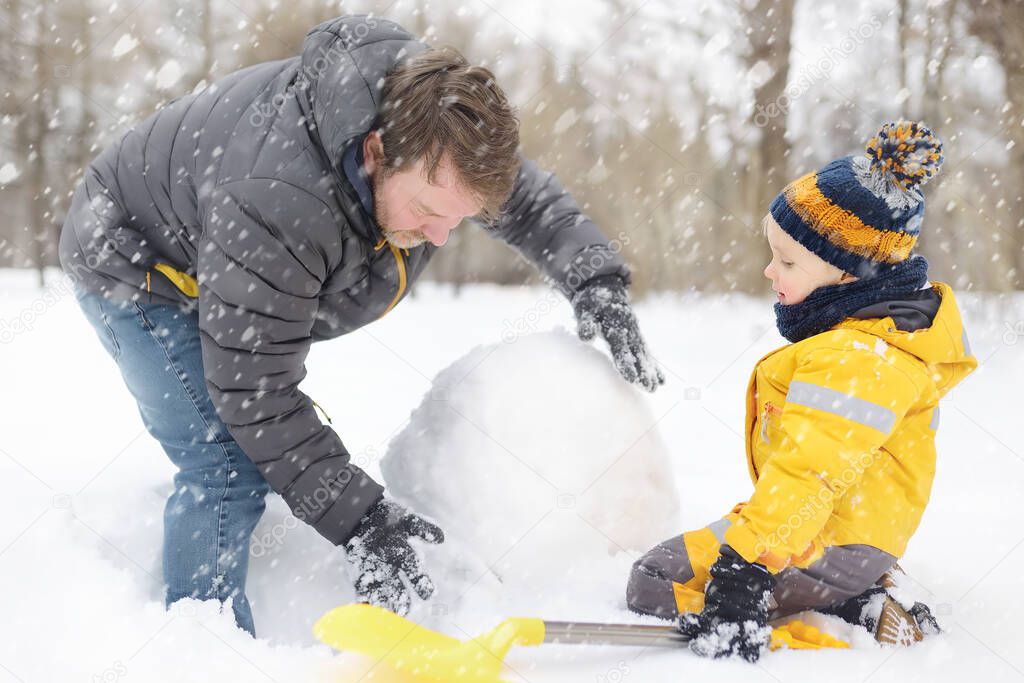 Little boy with his father building snowman in snowy park. Active outdoors leisure with children in winter. Kid during stroll in a snowy winter park