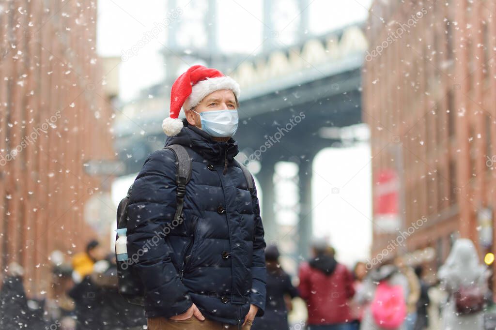 Middle aged man tourist wearing protective face mask is walking during a snowfall near Manhattan Bridge in New York on a snowy Christmas Eve. Winter Xmas holidays in NY. New Year vacations in NYC.