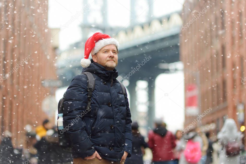 Middle aged man tourist in a Santa Claus hat is walking during a snowfall near Manhattan Bridge in New York on a snowy Christmas Eve. Winter Xmas holidays in NY. New Year vacations in NYC.