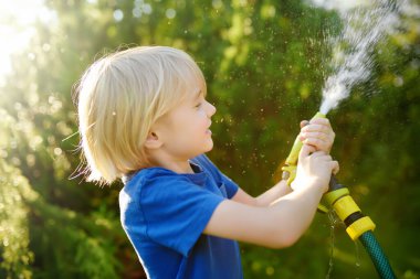 Çiçekleri sulayan, bahçe hortumuyla oynayan ve güneşli arka bahçede fıskiyeyle oynayan komik küçük çocuk. Anaokulu çocuğu su püskürterek eğleniyor. Çocuklar için yaz etkinliği.
