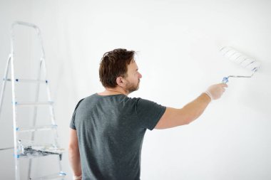 Evde tamirat yapan yakışıklı, olgun bir adamın portresi. Duvarı silindirle beyaza boyuyor. Kendiniz yapın. Diy. Elleriyle tamir ederler..