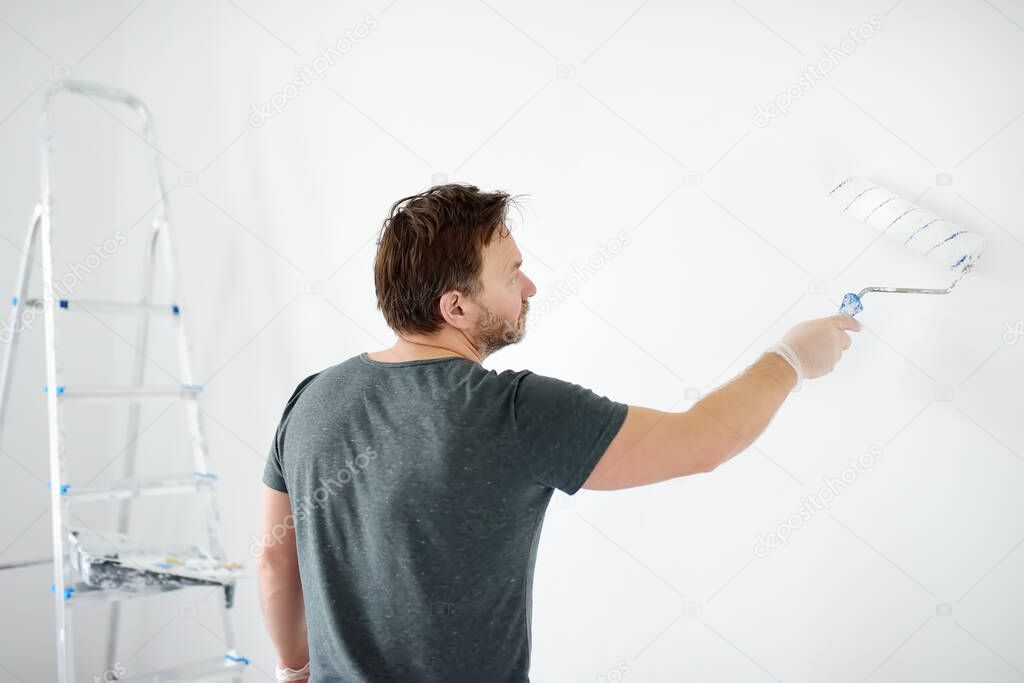 Portrait of a handsome mature man making repairs in the apartment. The person paints the wall white with roller. Do it yourself. DIY. Repair with their hands.
