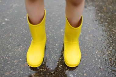 Küçük çocuk, yağmurlu yaz gününde küçük kasabada yürürken sarı lastik çizmeler giyiyor. Çocuk eğleniyor. Yağmurda çocuklar için açık hava oyunları.
