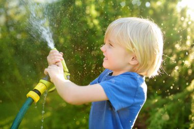 Çiçekleri sulayan, bahçe hortumuyla oynayan ve güneşli arka bahçede fıskiyeyle oynayan komik küçük çocuk. Anaokulu çocuğu su püskürterek eğleniyor. Çocuklar için yaz etkinliği.